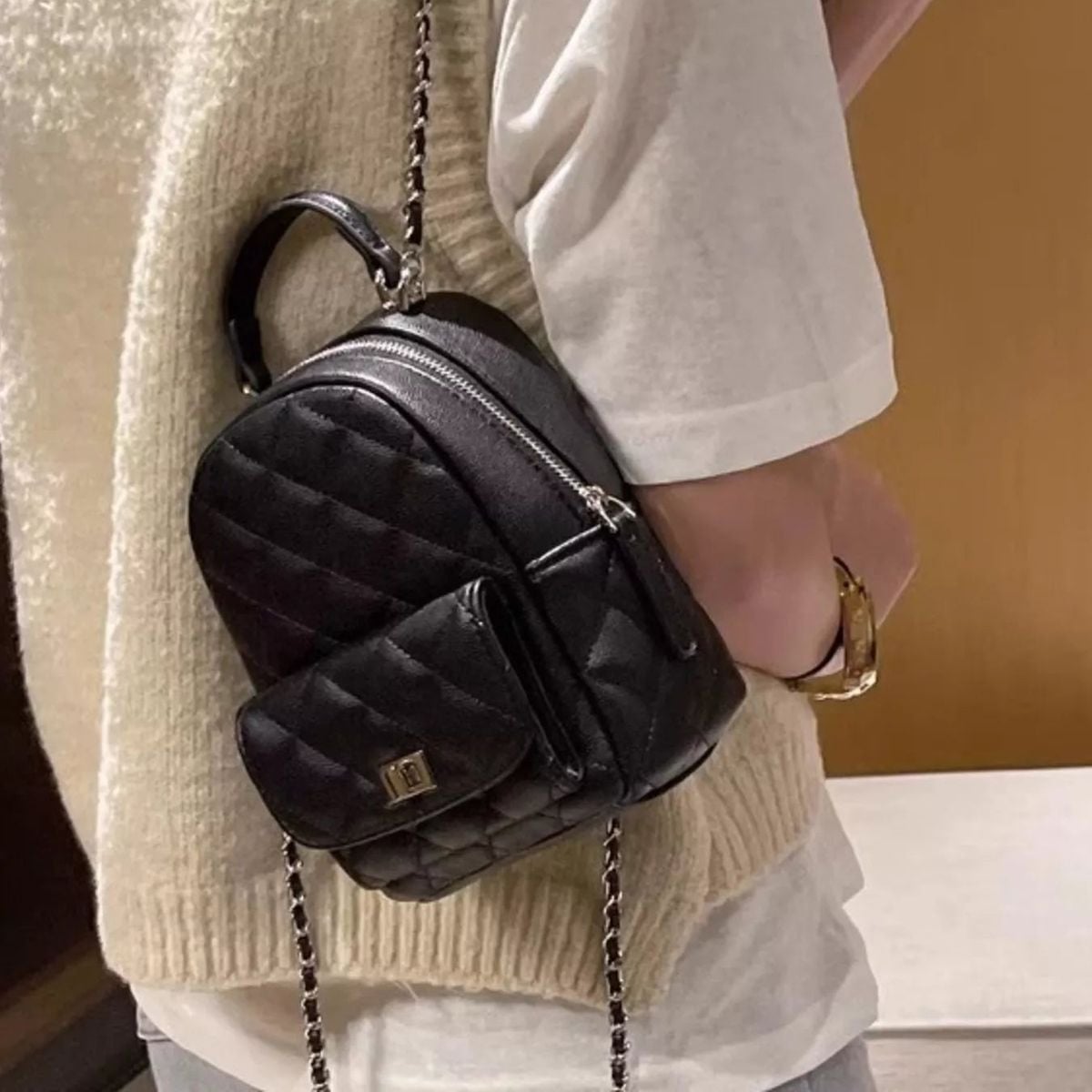 A person wearing a cream sweater and white shirt carries a small, quilted black leather backpack with a front pocket and chain straps over their shoulder.