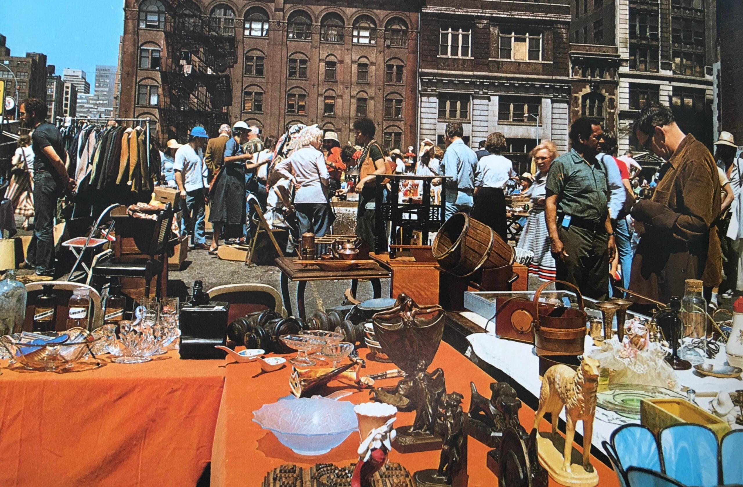 A busy outdoor flea market scene with people browsing tables filled with antiques, glassware, clothing, and various secondhand items, set against a backdrop of old brick buildings.