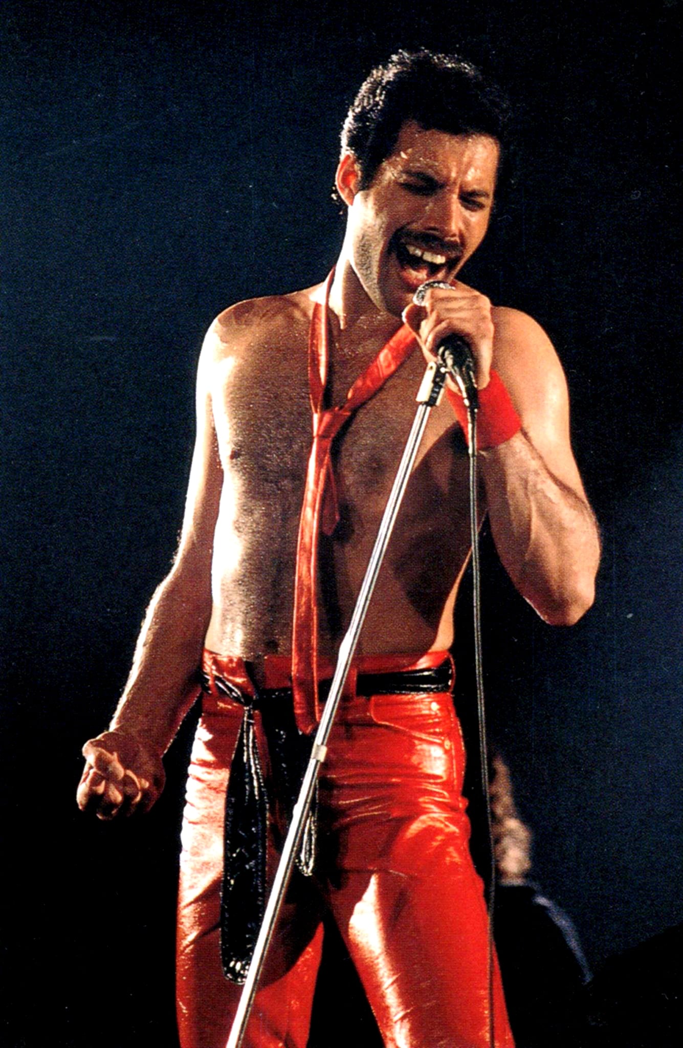 A man with short dark hair, a mustache, and a bare chest sings passionately into a microphone. He wears shiny red pants and a loosely tied red necktie, performing on stage under dramatic lighting.