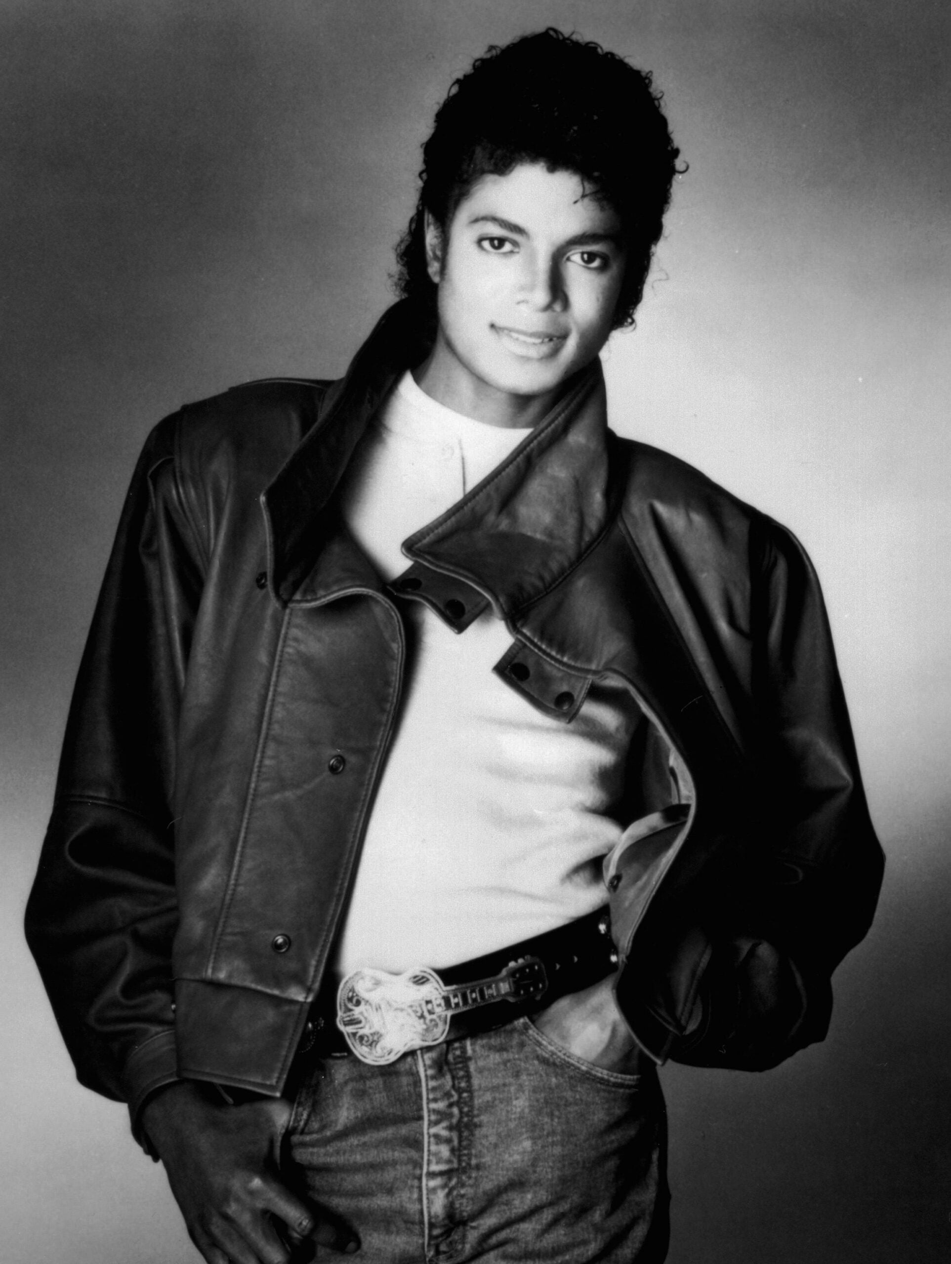 A young man with curly hair stands, smiling, wearing a leather jacket over a white shirt and jeans with a decorative belt. The photo is black and white with a plain background.