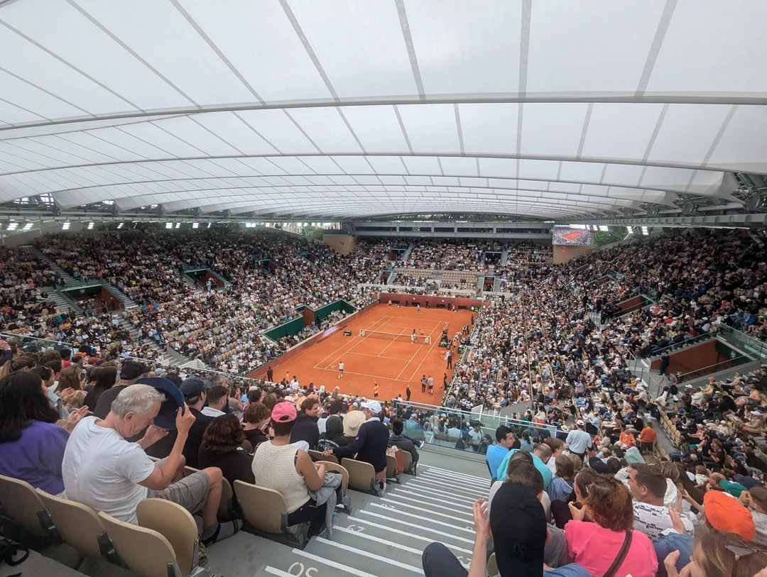 A packed stadium filled with spectators overlooks a clay tennis court where a doubles match is being played under a large, translucent roof. The crowd is engaged, and the atmosphere is lively.