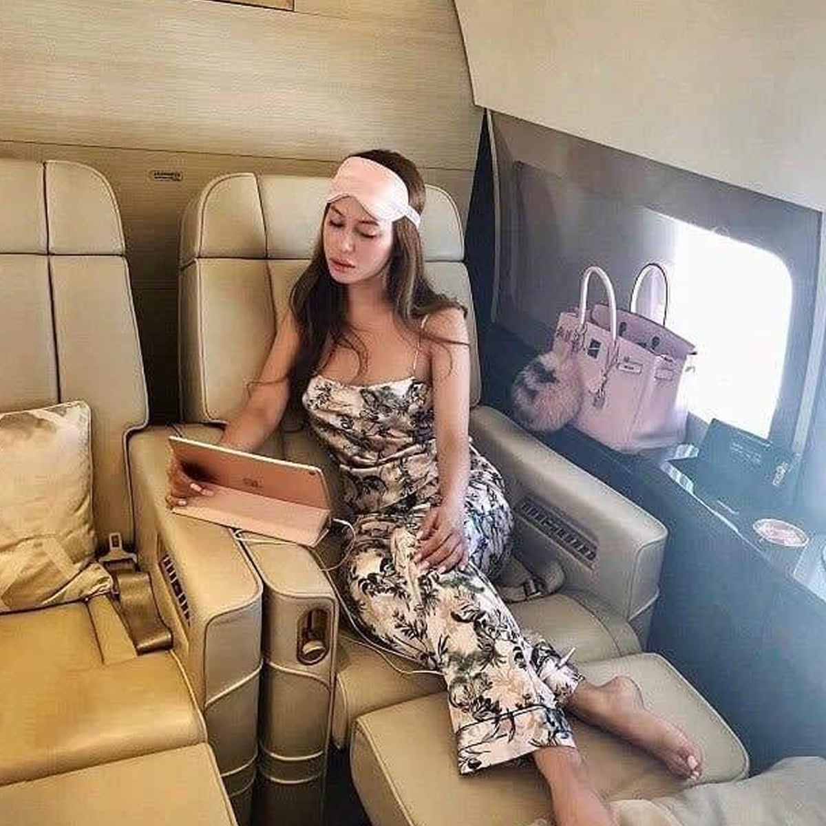 A woman in a floral jumpsuit and pink sleep mask sits in a luxurious airplane seat, looking at a tablet. A pink designer handbag rests on the seat next to her, and she appears relaxed during the flight.