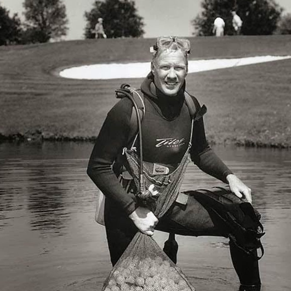 A man in a wetsuit and snorkel stands knee-deep in a pond, holding a mesh bag full of golf balls, with a golf course and sand trap in the background.