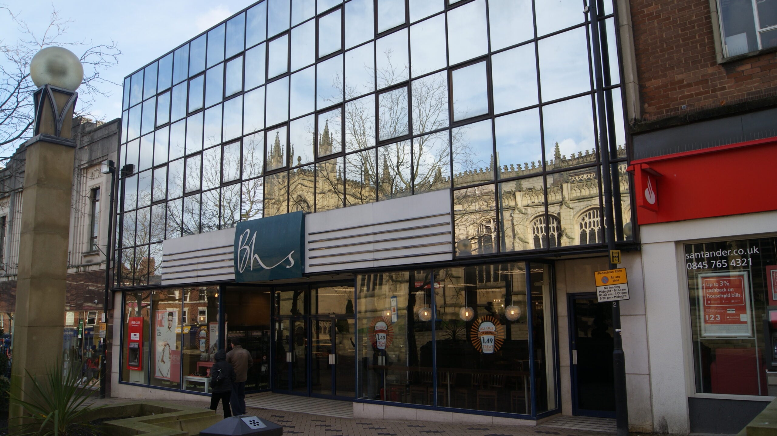 A modern glass-fronted building with the "BHS" sign, reflecting a historic church and trees. People walk by the entrance, and a Santander bank is visible on the right side.