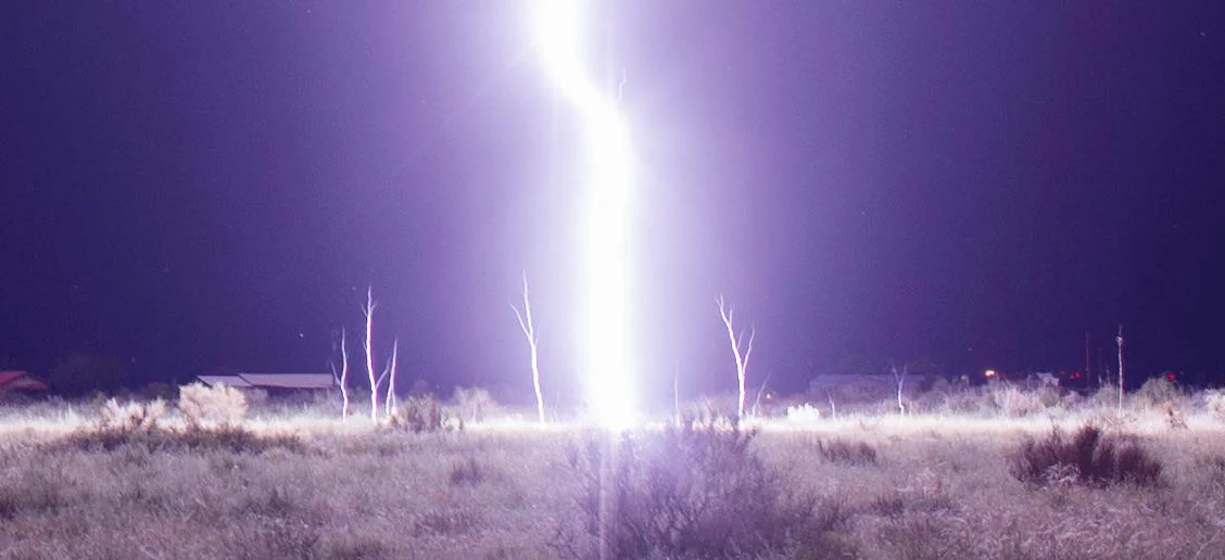 A bright bolt of lightning strikes the ground in an open field at night, illuminating the landscape and nearby leafless trees under a dark sky.