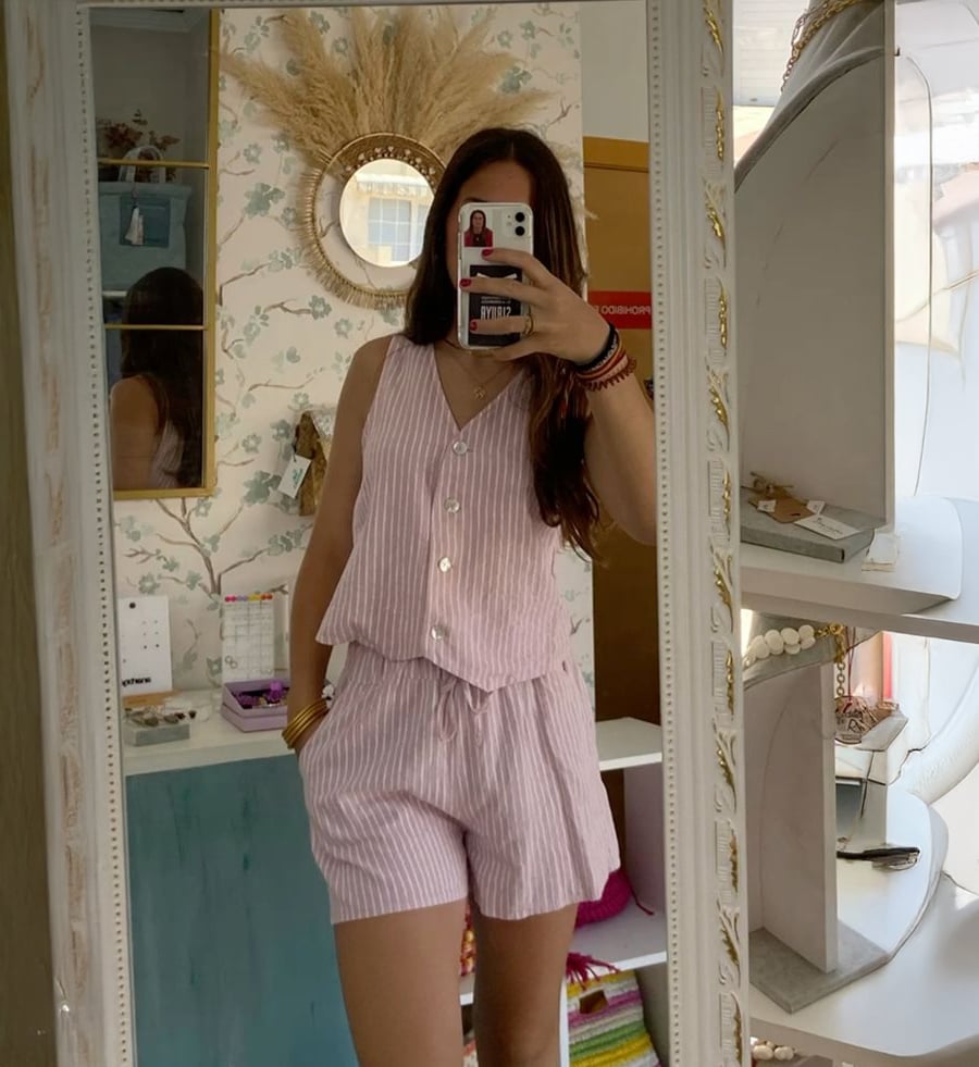 A person wearing light pink and white striped pajamas takes a mirror selfie in a cozy room with floral wallpaper, shelves, and decor items. Their face is partially covered by their phone.