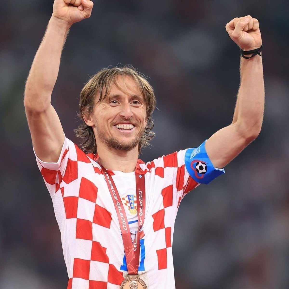 A smiling soccer player wearing a red and white checkered jersey and captain’s armband raises both fists in celebration, with a bronze medal around his neck.