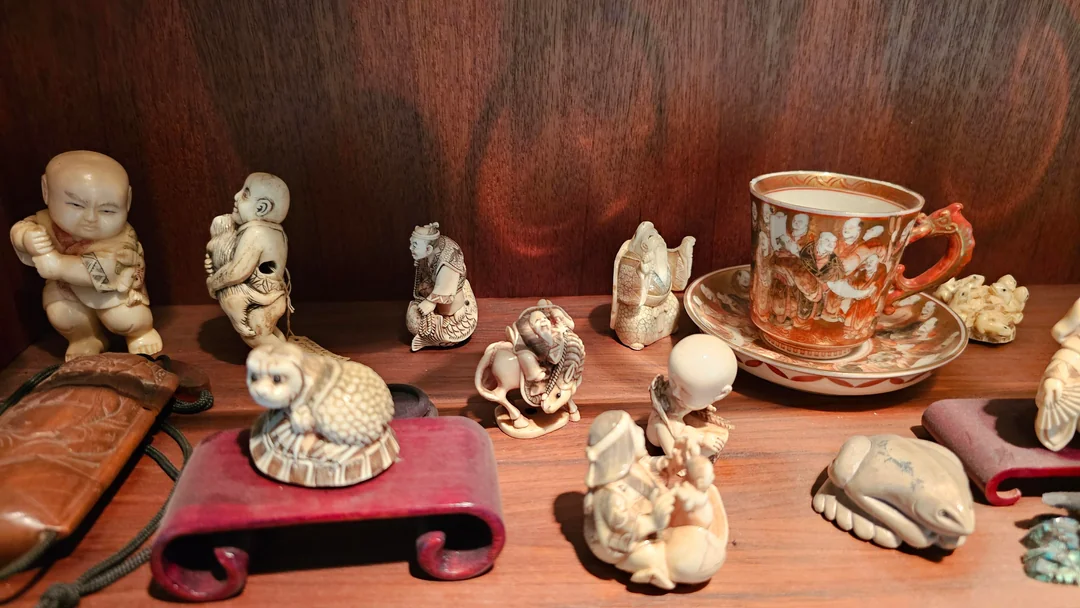 A collection of small ivory carvings and figurines is displayed on a wooden shelf, alongside an ornate teacup and saucer with detailed designs.