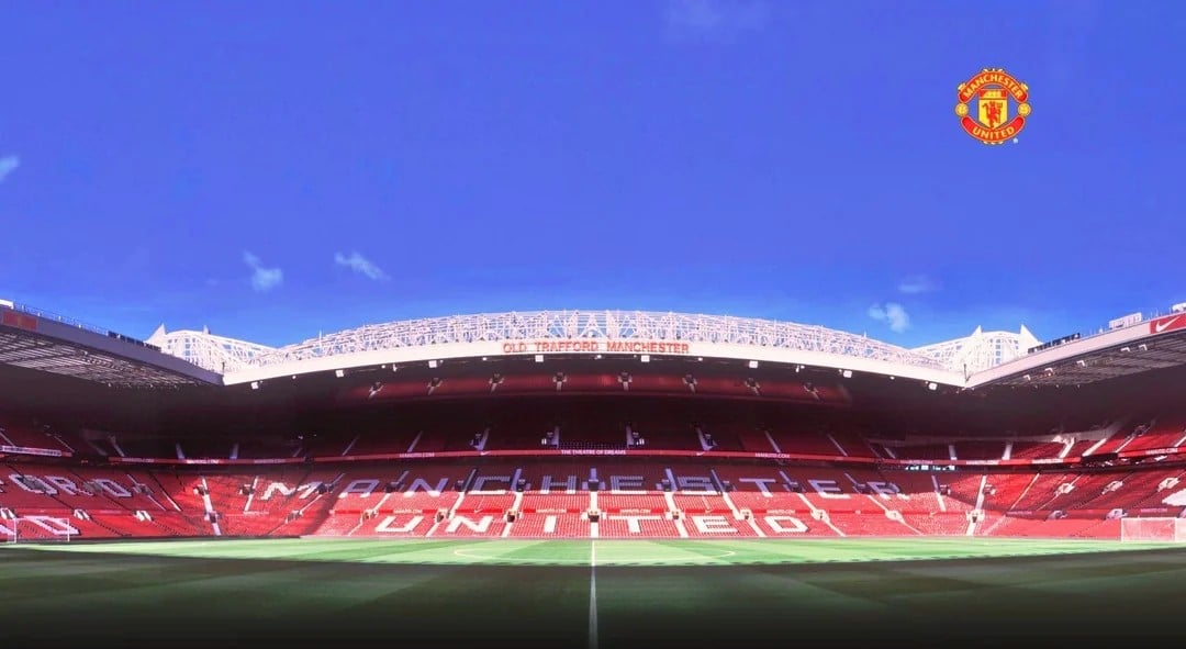 Wide-angle view of an empty Old Trafford football stadium, home to Manchester United, with the club’s logo in the top right corner and clear blue sky above the stands.
