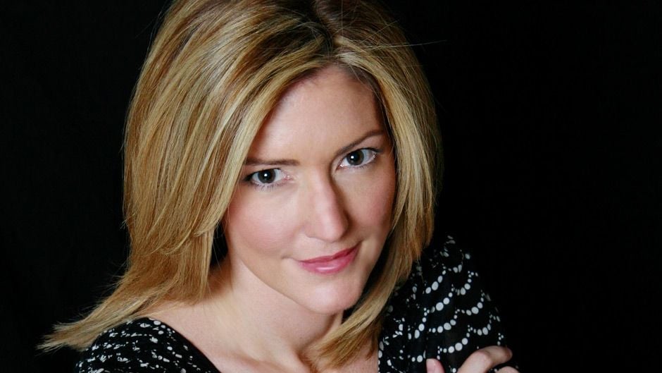 A woman with straight, shoulder-length blonde hair and fair skin looks at the camera. She is wearing a black patterned top and is set against a dark background.