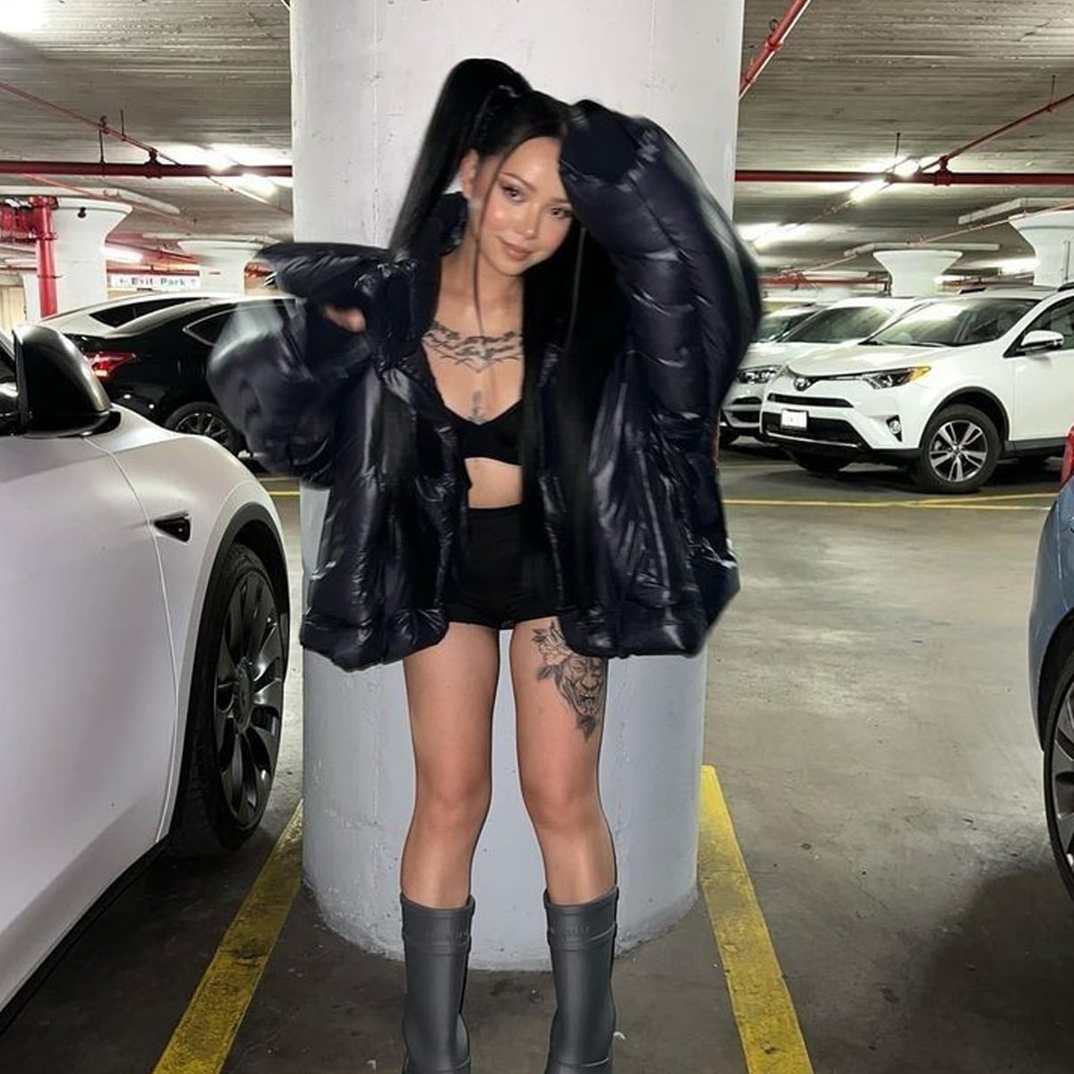 A woman with long black hair in pigtails poses in a parking garage, wearing a black puffer jacket, black crop top, shorts, and gray boots. She has tattoos on her thigh and a necklace with chunky silver chains.