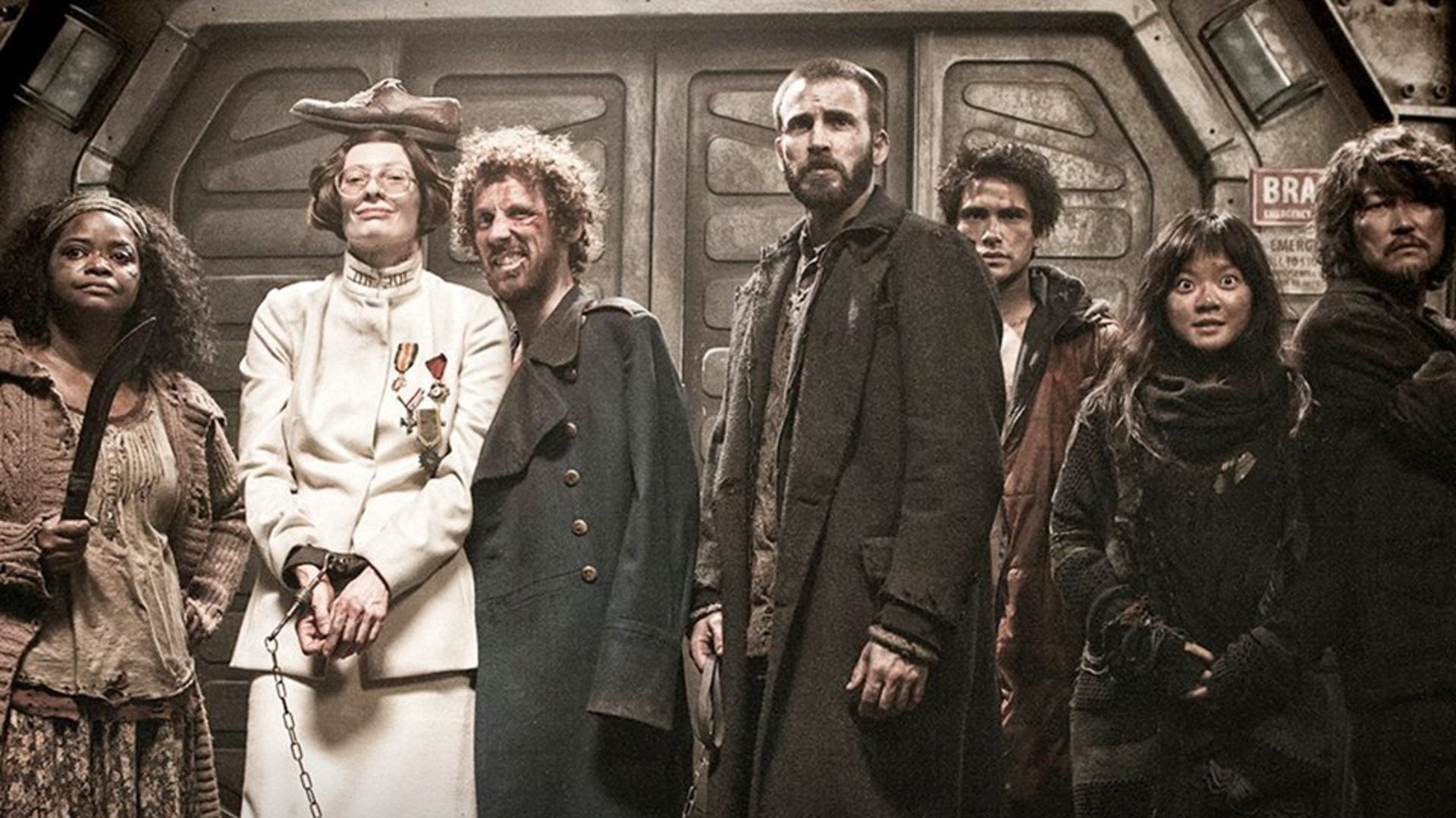 A group of seven people in worn and mismatched clothing stand in a dimly lit, industrial train car. One woman in white stands out, smiling with a shoe on her head, while others have serious or curious expressions.