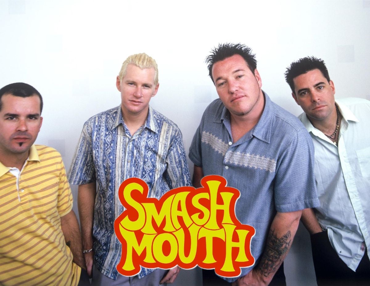 Four men stand close together against a plain backdrop, with the colorful "Smash Mouth" band logo in front of them. They wear casual shirts and face the camera with relaxed expressions.