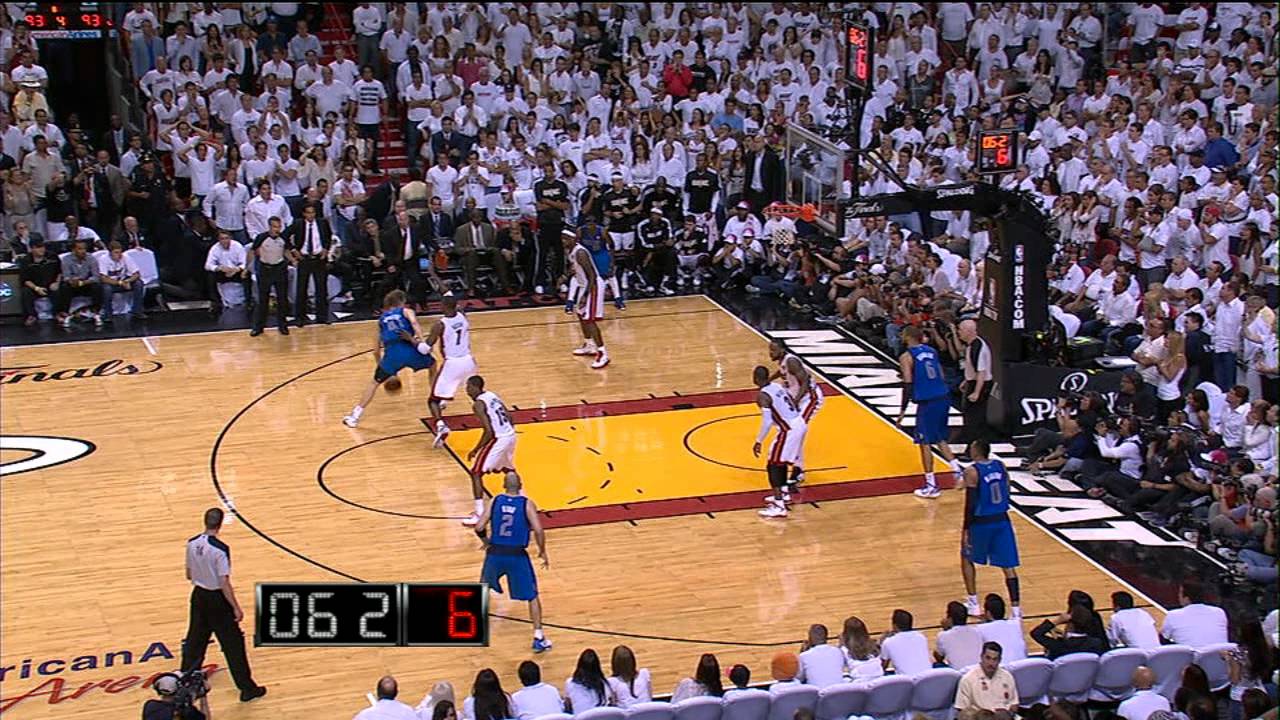 A basketball game is in progress, with one team in blue uniforms preparing to shoot while the opposing team in white defends. The scoreboard shows 62–61 and the shot clock reads 6 seconds. The arena is packed with fans.