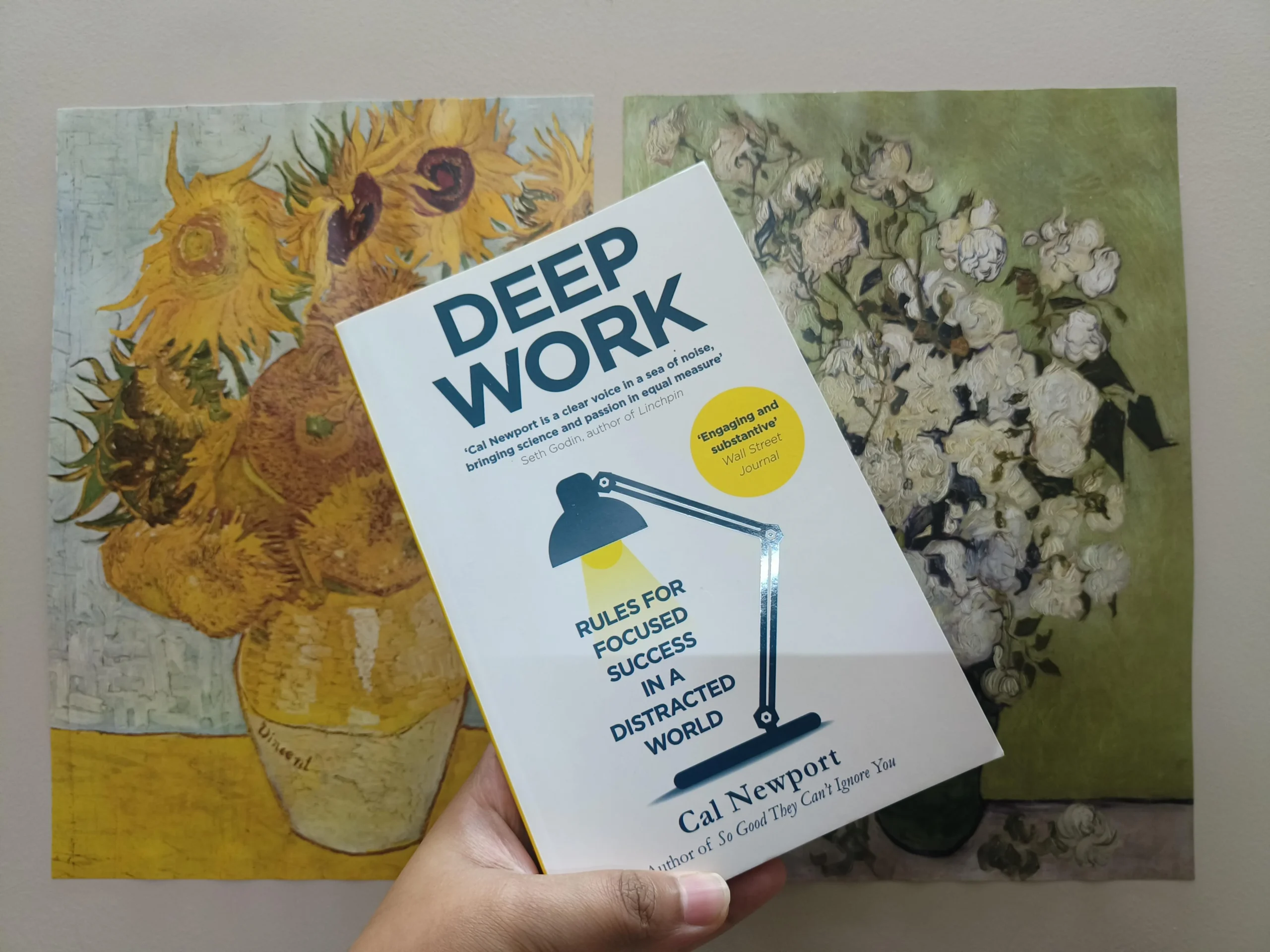 A hand holds the book "Deep Work" by Cal Newport in front of two paintings featuring flower arrangements, one with sunflowers and the other with white blossoms, both in vases.