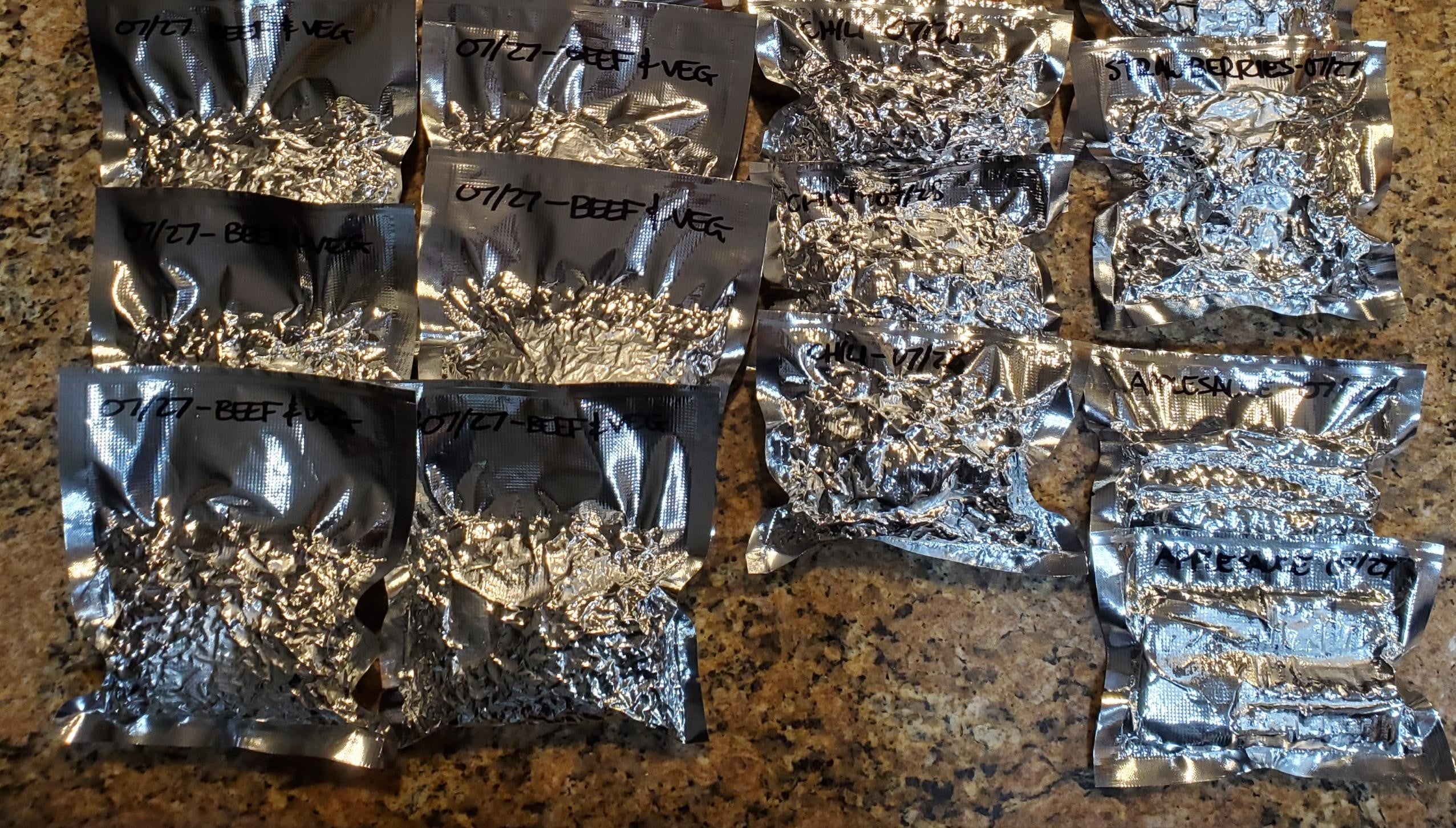 Several vacuum-sealed silver pouches labeled "07/27 - BEEF VEG" and other text are arranged on a granite countertop. The contents appear to be food portions, and the labels indicate the package dates and possibly meal types.
