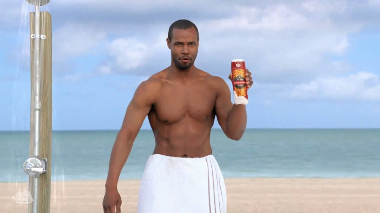 A shirtless man with a towel wrapped around his waist stands on a beach near a shower, holding up a bottle of Old Spice body wash, with the ocean and cloudy sky in the background.