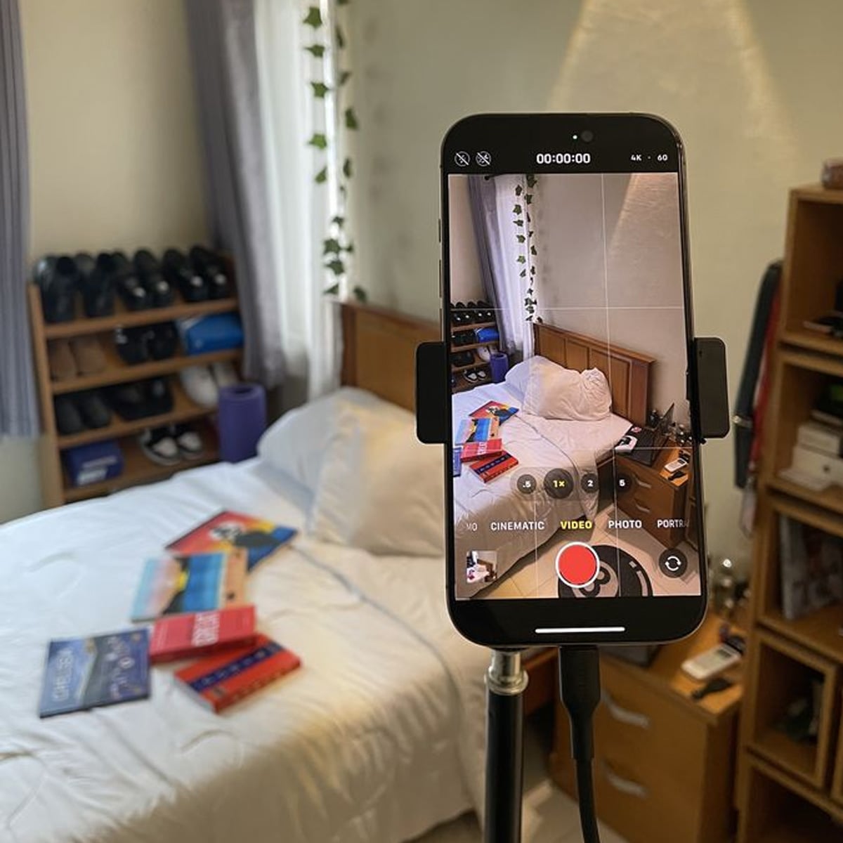 A smartphone on a tripod records a video of a tidy bedroom with a made bed, books scattered on the duvet, shelves with shoes, and bookshelves against the walls.