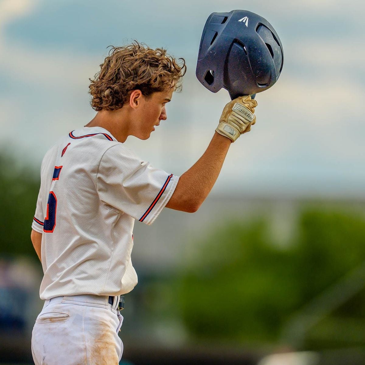 A young baseball player in a white uniform holds his helmet up in one hand, looking off to the side. Trees and a blurred background suggest an outdoor field setting.