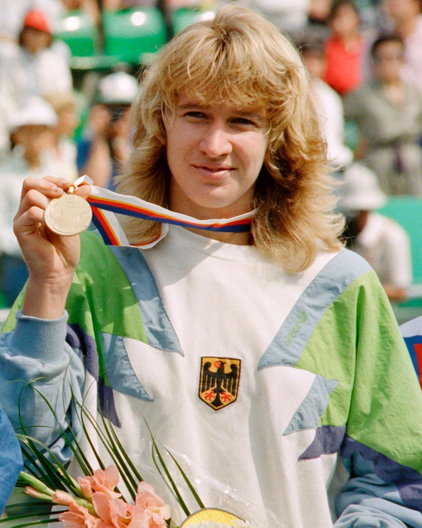 A young athlete with blonde hair holds up a gold medal and wears a sweatshirt with the German flag emblem. There are flowers in the foreground and a crowd in the background.