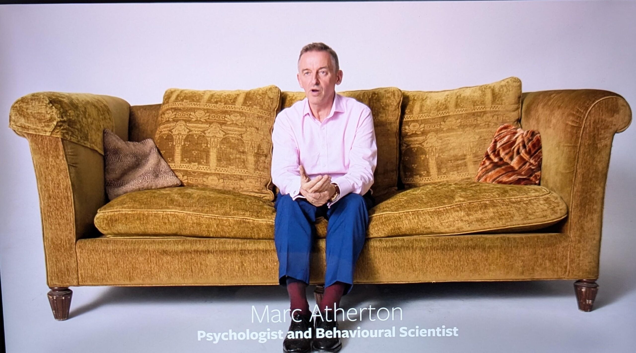 A man in a pink shirt and blue pants sits on a mustard yellow sofa with patterned cushions. Text at the bottom reads: "Marc Atherton, Psychologist and Behavioural Scientist.
