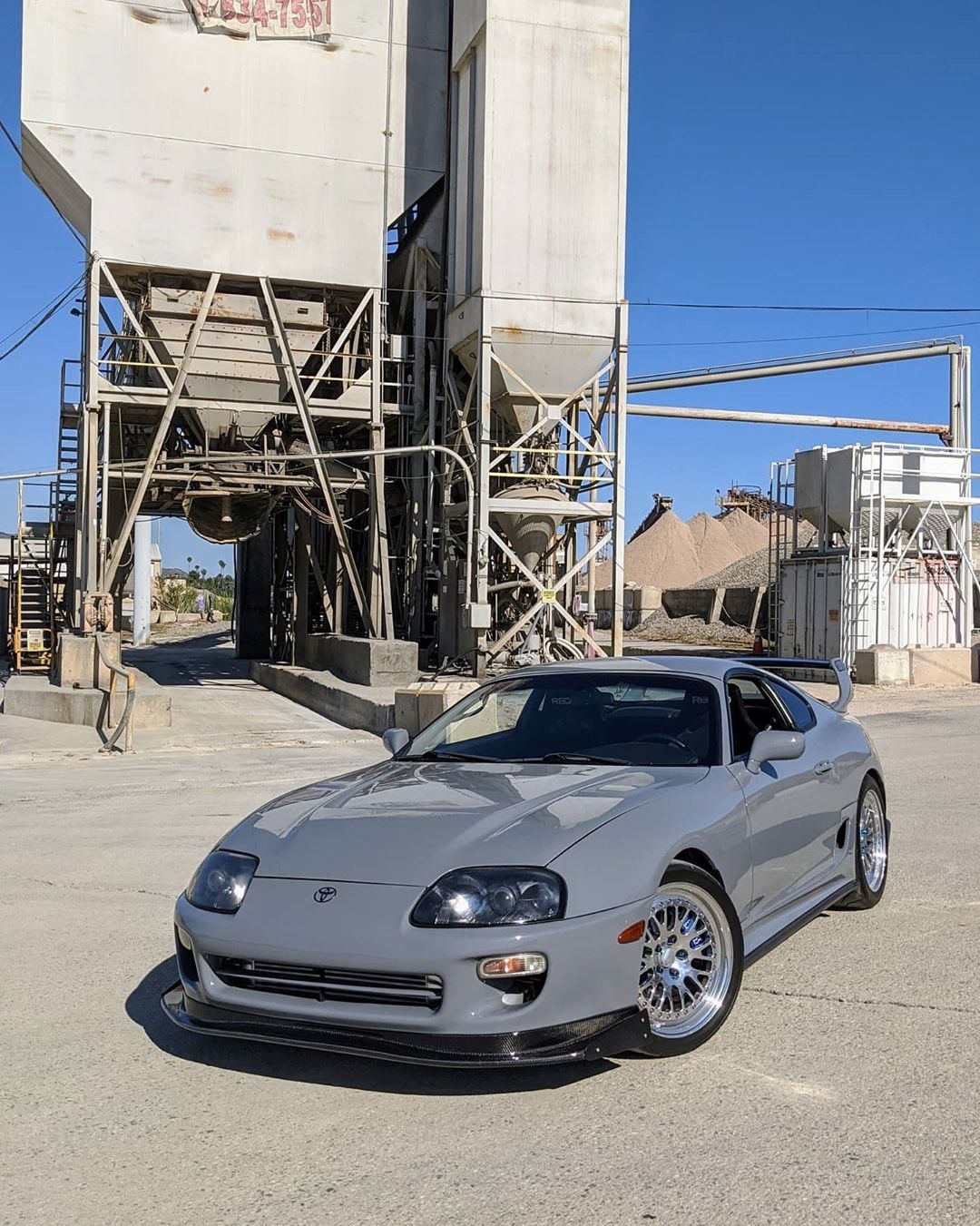 A silver Toyota Supra sports car is parked on a sunny day in front of an industrial concrete plant with metal structures and sand piles in the background.
