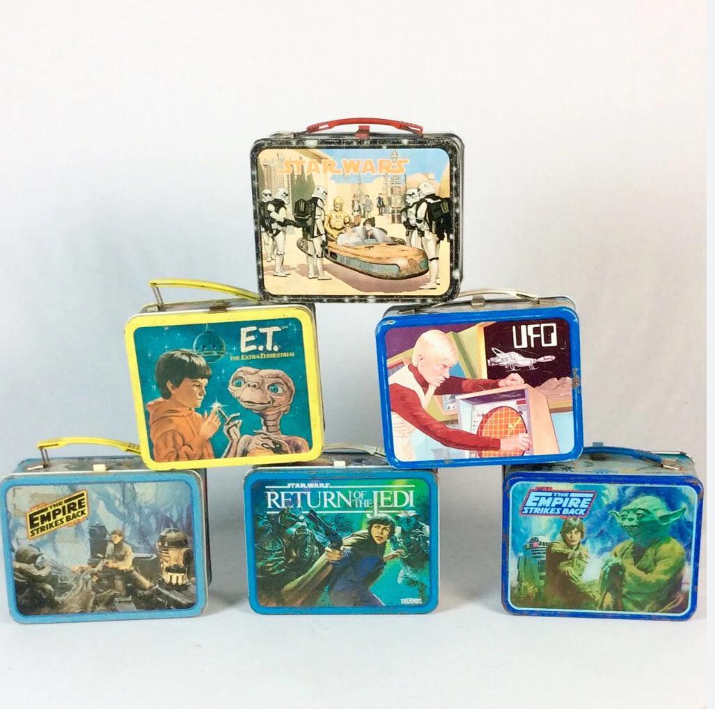 Six vintage metal lunchboxes are stacked in a pyramid, featuring designs from Star Wars, E.T., UFO, Return of the Jedi, and The Empire Strikes Back with colorful artwork and characters.
