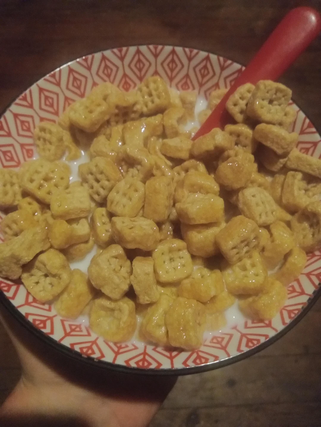 A colorful bowl filled with cereal squares and milk, with a red spoon placed inside. The bowl has a red geometric pattern on the rim and is being held in one hand.