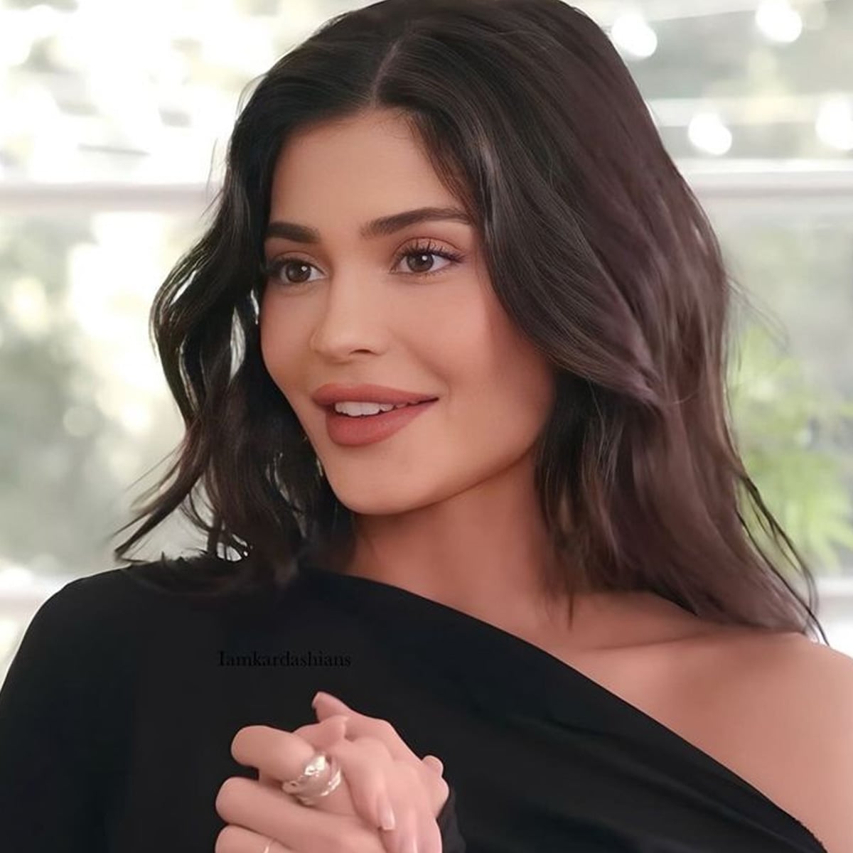 A woman with wavy dark hair and natural makeup smiles softly, wearing an off-the-shoulder black top. The background is softly blurred with hints of greenery and string lights.