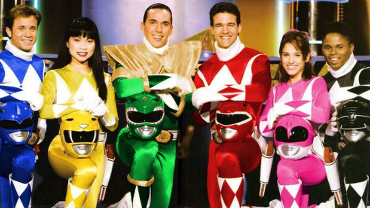 Six people dressed as Power Rangers in colorful suits—blue, yellow, green, red, pink, and black—pose and smile together, each holding their matching helmets in front of them against a bright background.
