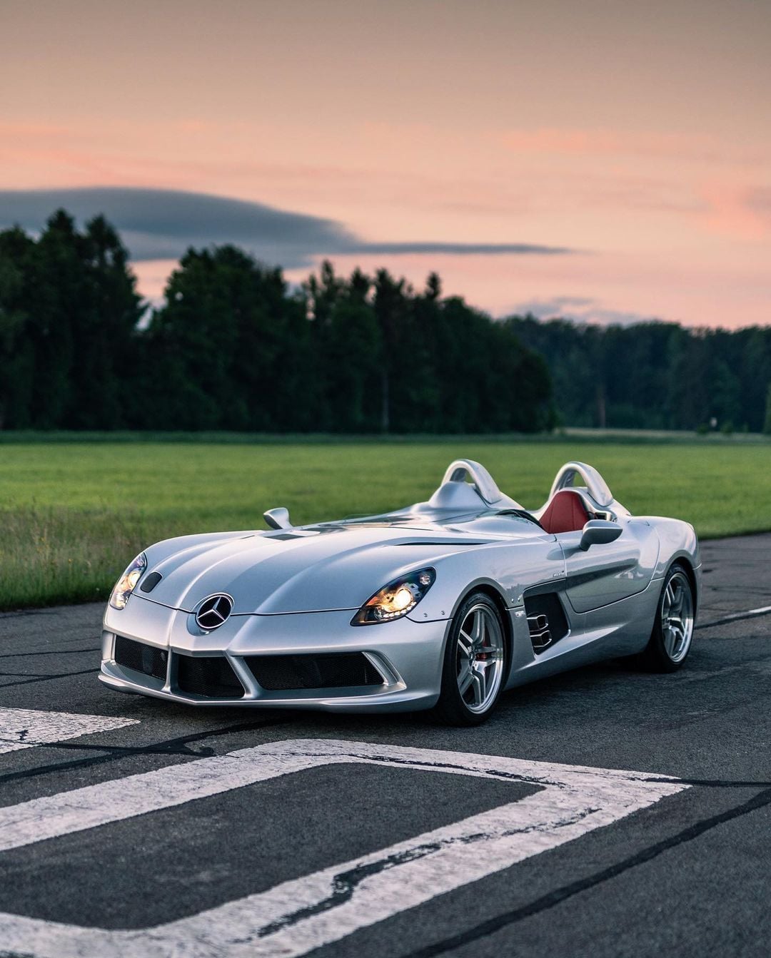 A silver Mercedes-Benz SLR Stirling Moss with an open cockpit is parked on a paved road at sunset, with green grass and trees in the background.