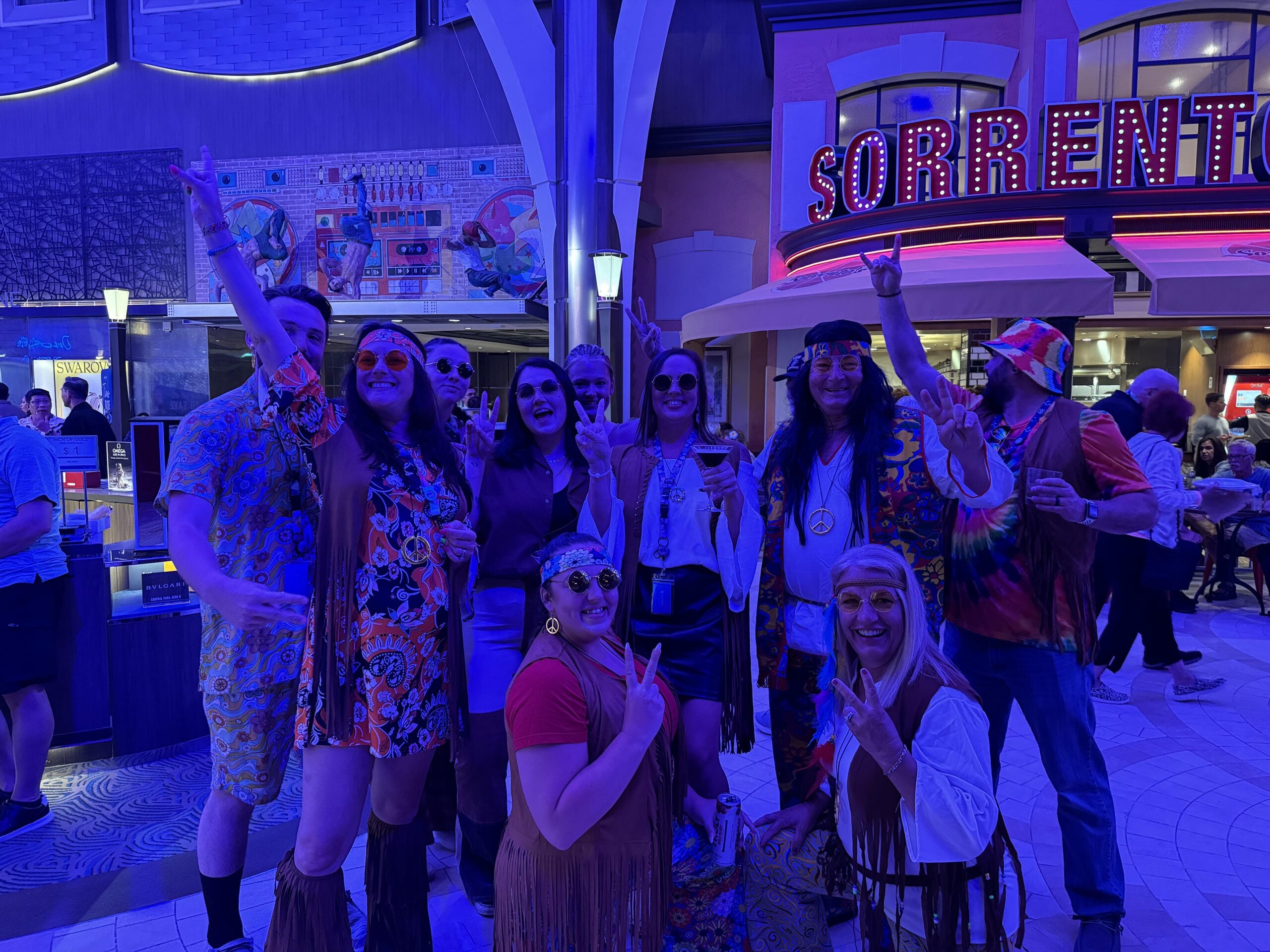 A group of people in colorful 1960s hippie costumes pose together indoors, smiling and making peace signs, under bright purple lighting near a “Sorrento’s” sign.