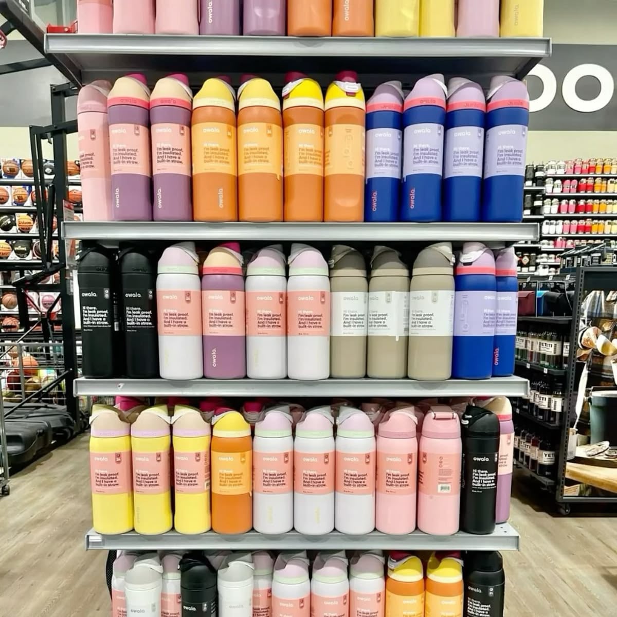 Shelves in a store display rows of colorful insulated water bottles in gradient shades of pink, orange, yellow, blue, gray, and black, neatly arranged by color. Sports equipment is visible in the background.
