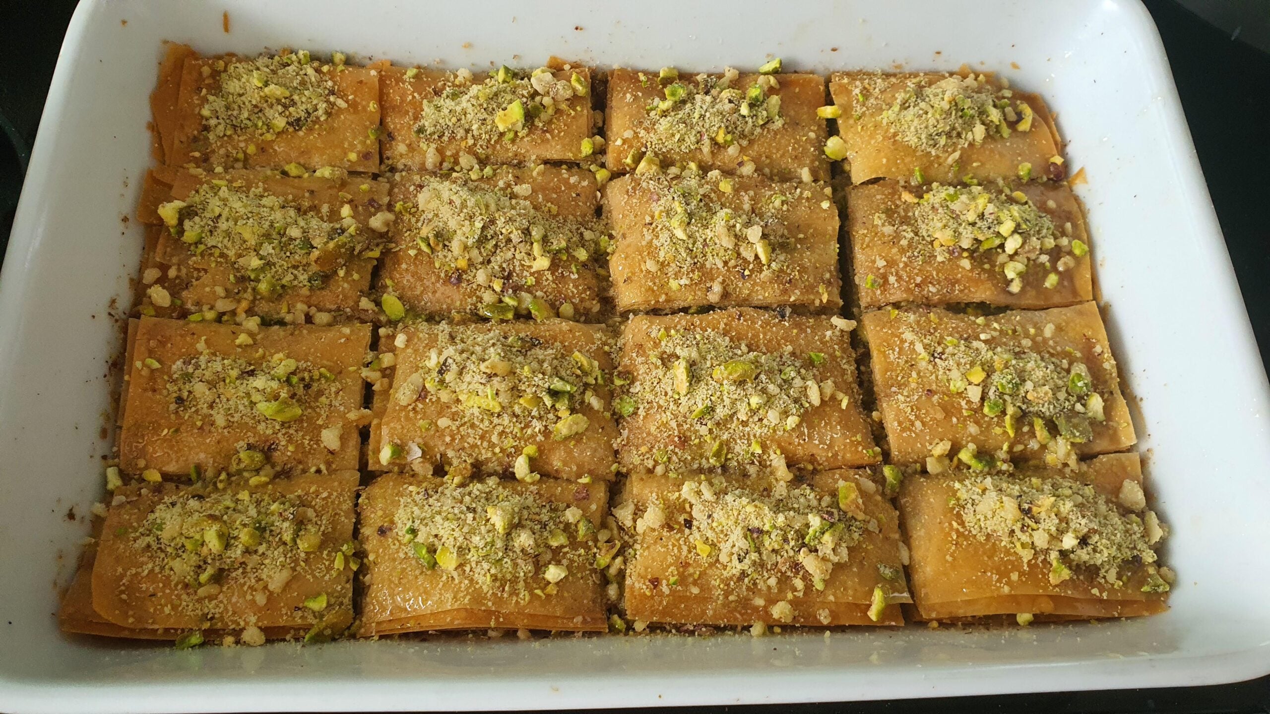 A tray of baklava slices topped with chopped pistachios, arranged neatly in rows within a white rectangular baking dish.