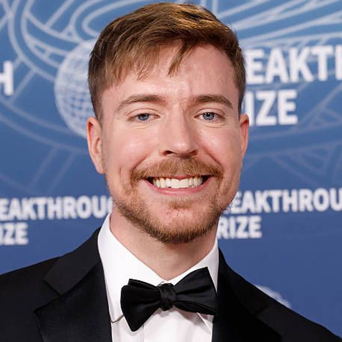 A man in a black tuxedo and bow tie smiles in front of a blue backdrop with "BREAKTHROUGH PRIZE" written on it.