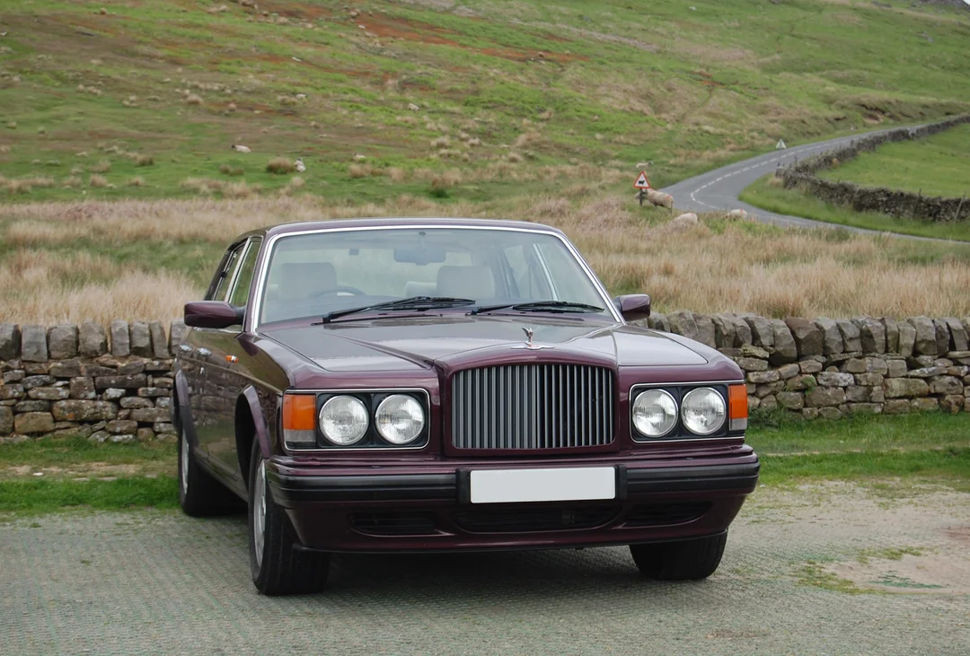 A vintage maroon luxury car is parked on a grassy area in front of a stone wall, with rolling green hills and a winding road in the background.
