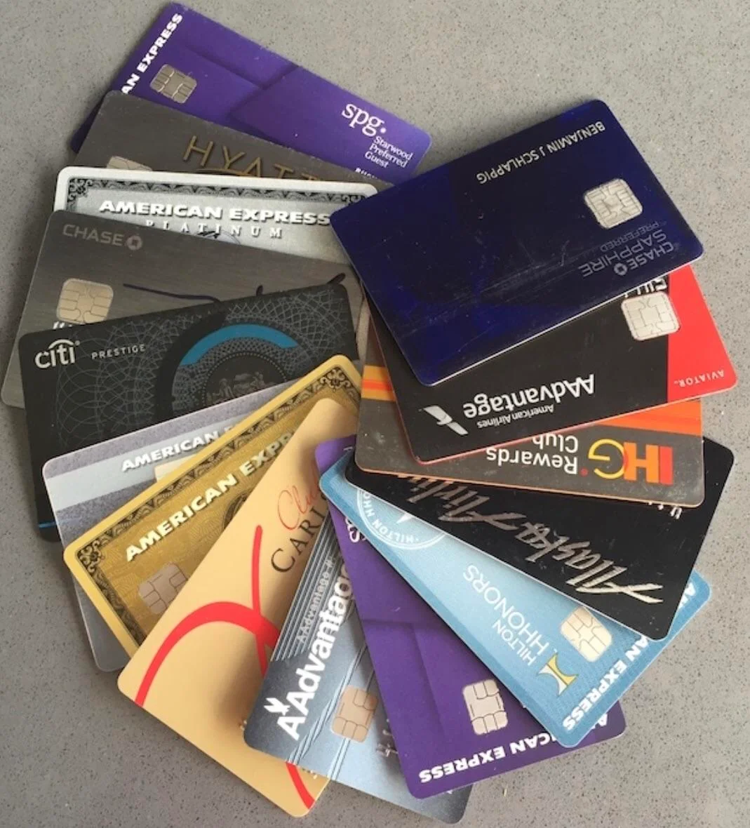 A scattered pile of various credit cards and rewards cards from different banks and travel programs lies on a gray surface. The cards feature different colors and logos, including American Express, Chase, Citi, and Hilton.