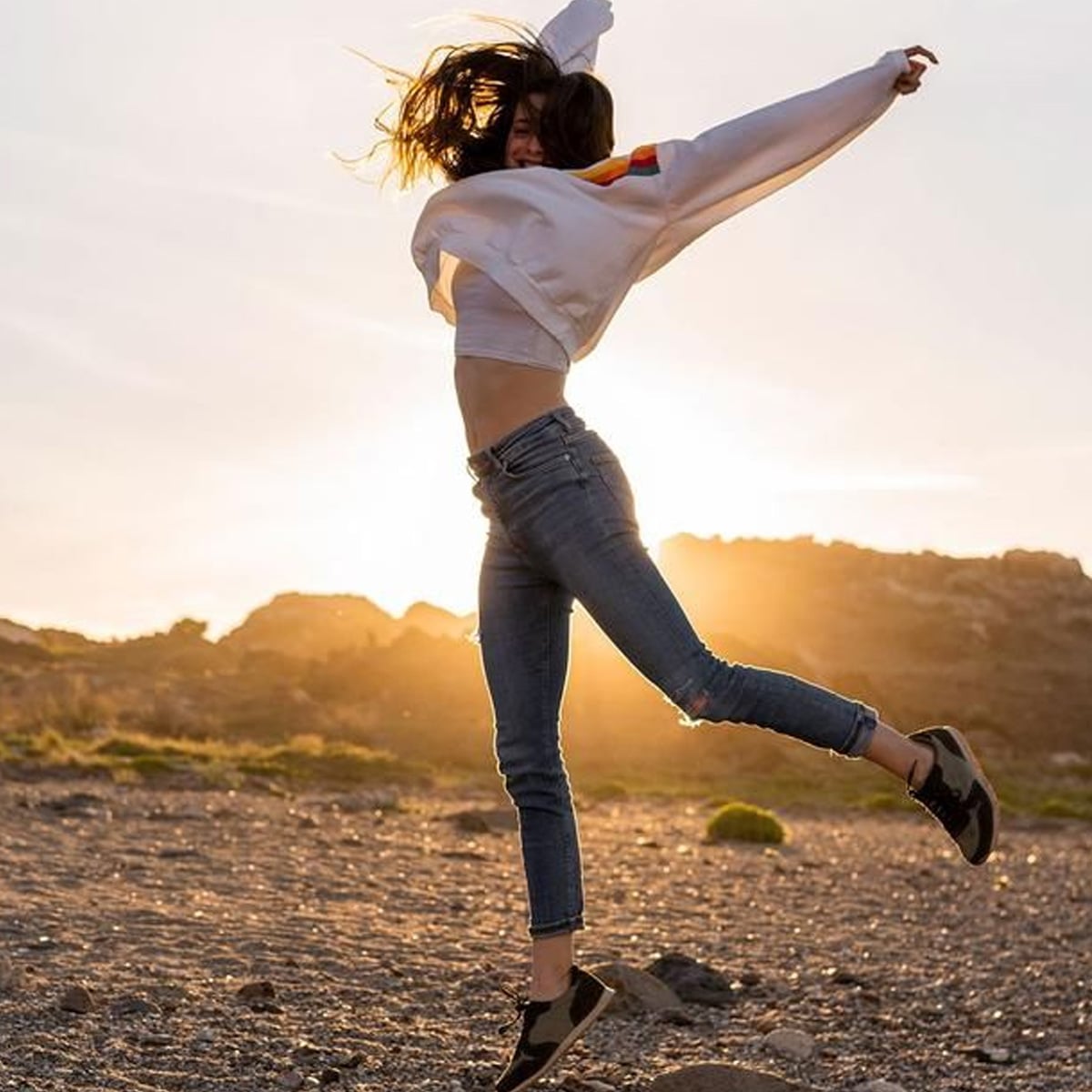 A person wearing jeans, sneakers, and a light sweater jumps joyfully on a rocky landscape at sunset, with the sun casting a warm glow behind them.