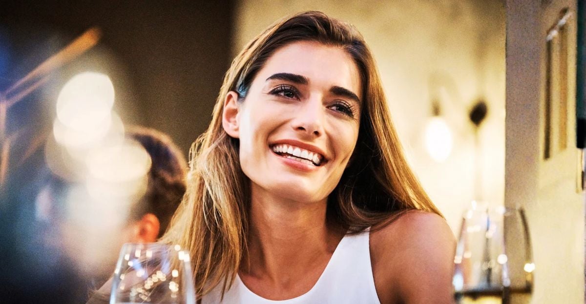 A woman with long brown hair smiles brightly in a warmly lit indoor setting, wearing a sleeveless white top. There are glasses and blurred lights in the foreground, creating a cheerful atmosphere.