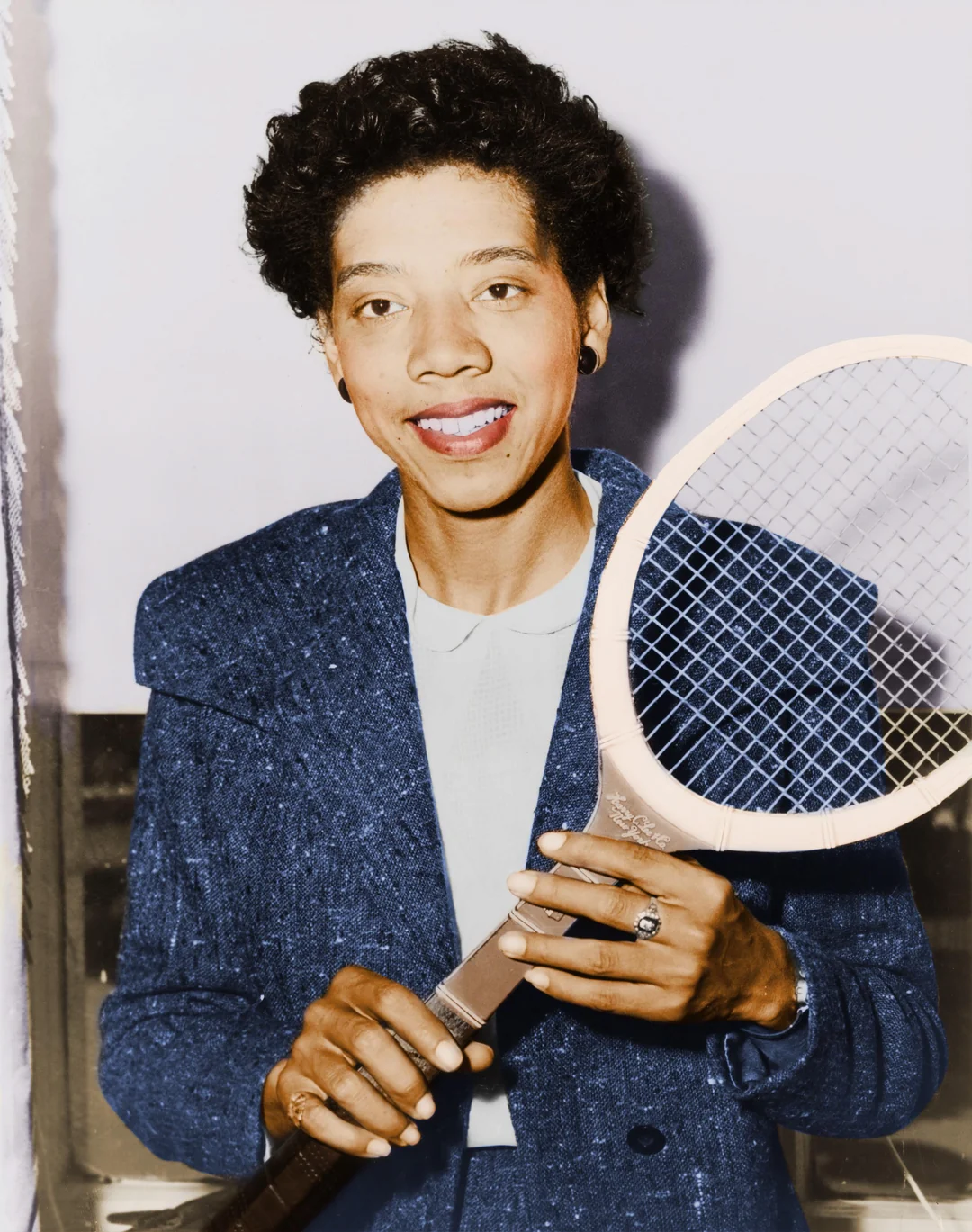 A smiling woman wearing a blue suit jacket holds a wooden tennis racket upright with both hands, standing against a neutral background.
