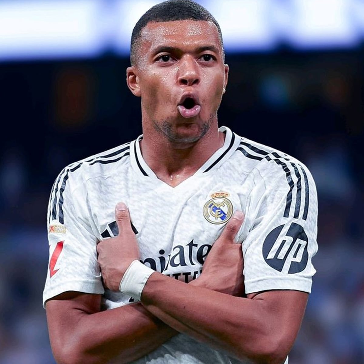 A soccer player wearing a white Real Madrid jersey celebrates with arms crossed over his chest, mouth open in excitement, on a blurred stadium background.