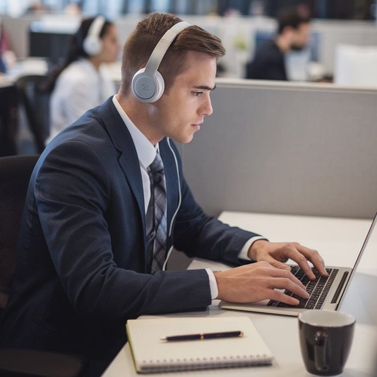 A man in a suit wearing headphones works on a laptop at an office desk. A notebook and a coffee mug are on the desk. Other people are working in the background.