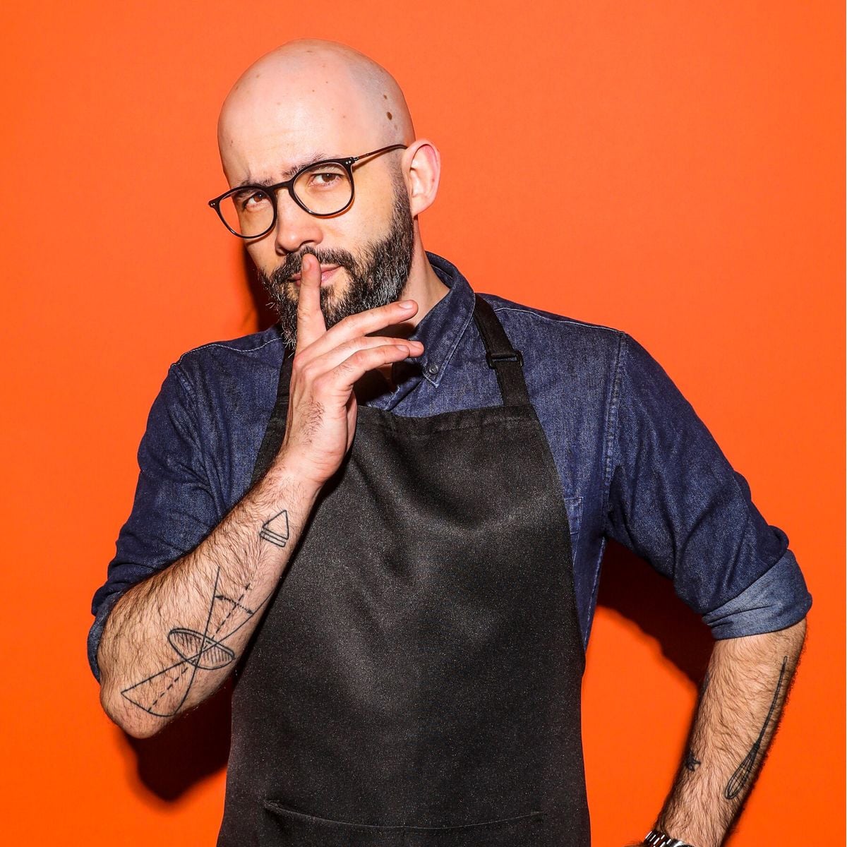 A bald man with glasses and tattoos, wearing a denim shirt and black apron, stands against an orange background, holding a finger to his lips in a "shh" gesture.