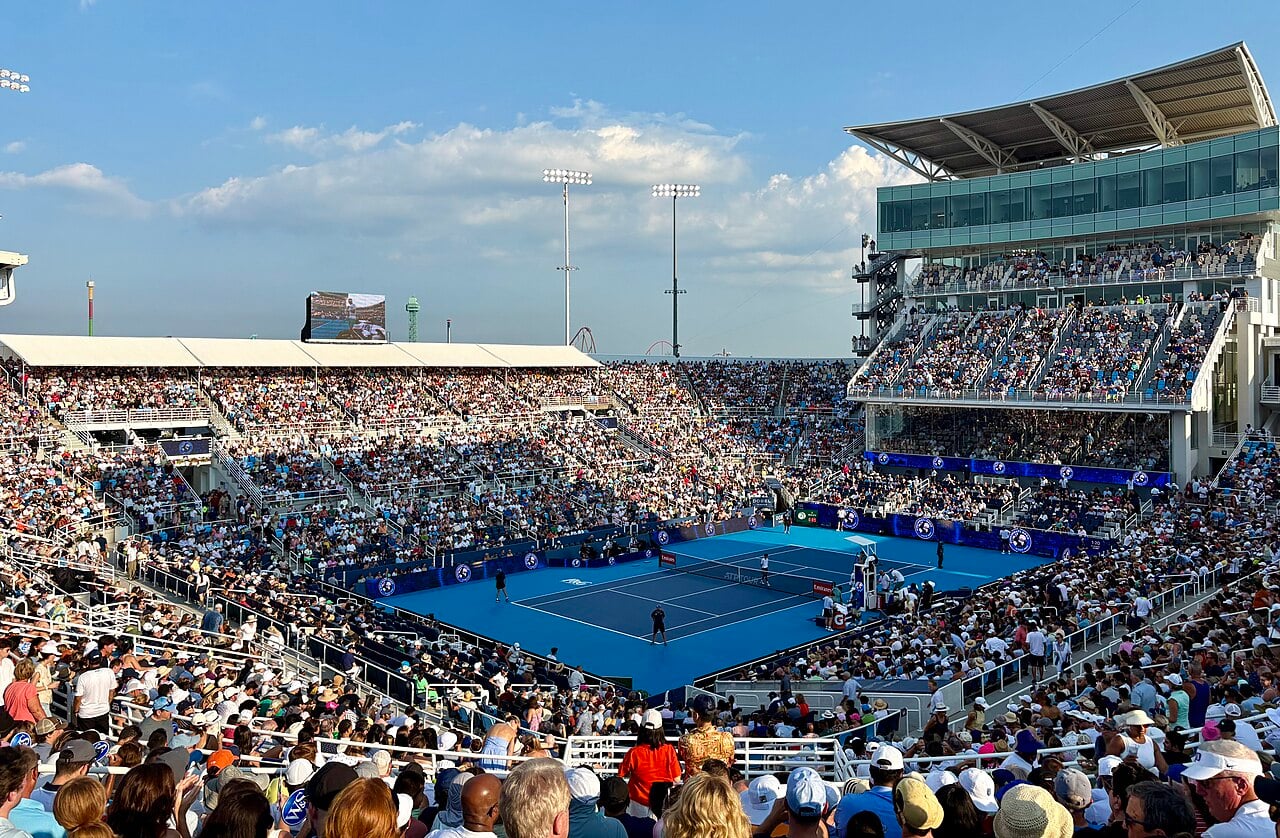 A packed outdoor tennis stadium with spectators watching a match on a blue hard court under a clear sky, with tall lights and a large video screen visible in the background.