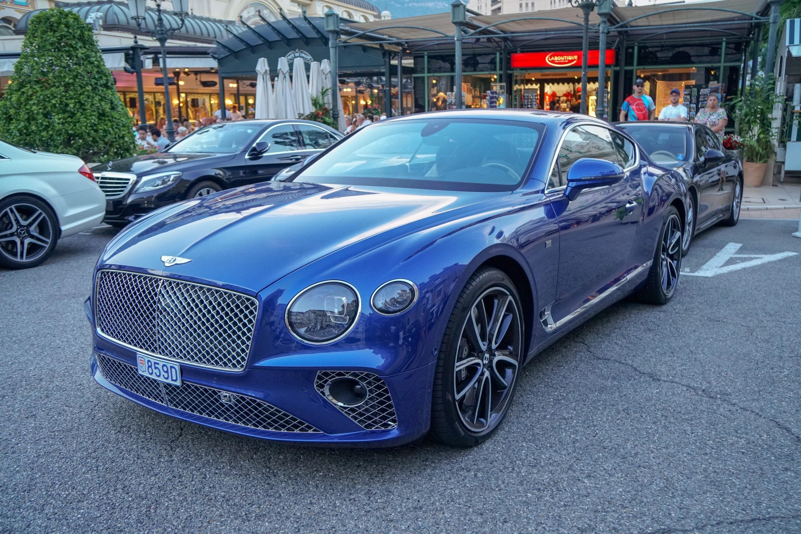 A blue Bentley Continental GT is parked on a street in front of shops and restaurants, with other cars and people in the background.