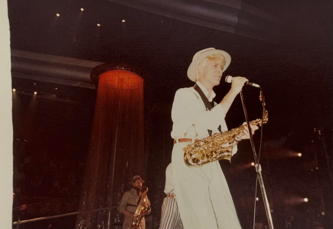 A musician in a white outfit and hat holds a saxophone and sings into a microphone on stage. Another musician with a saxophone is in the background, with stage lights and curtains behind them.