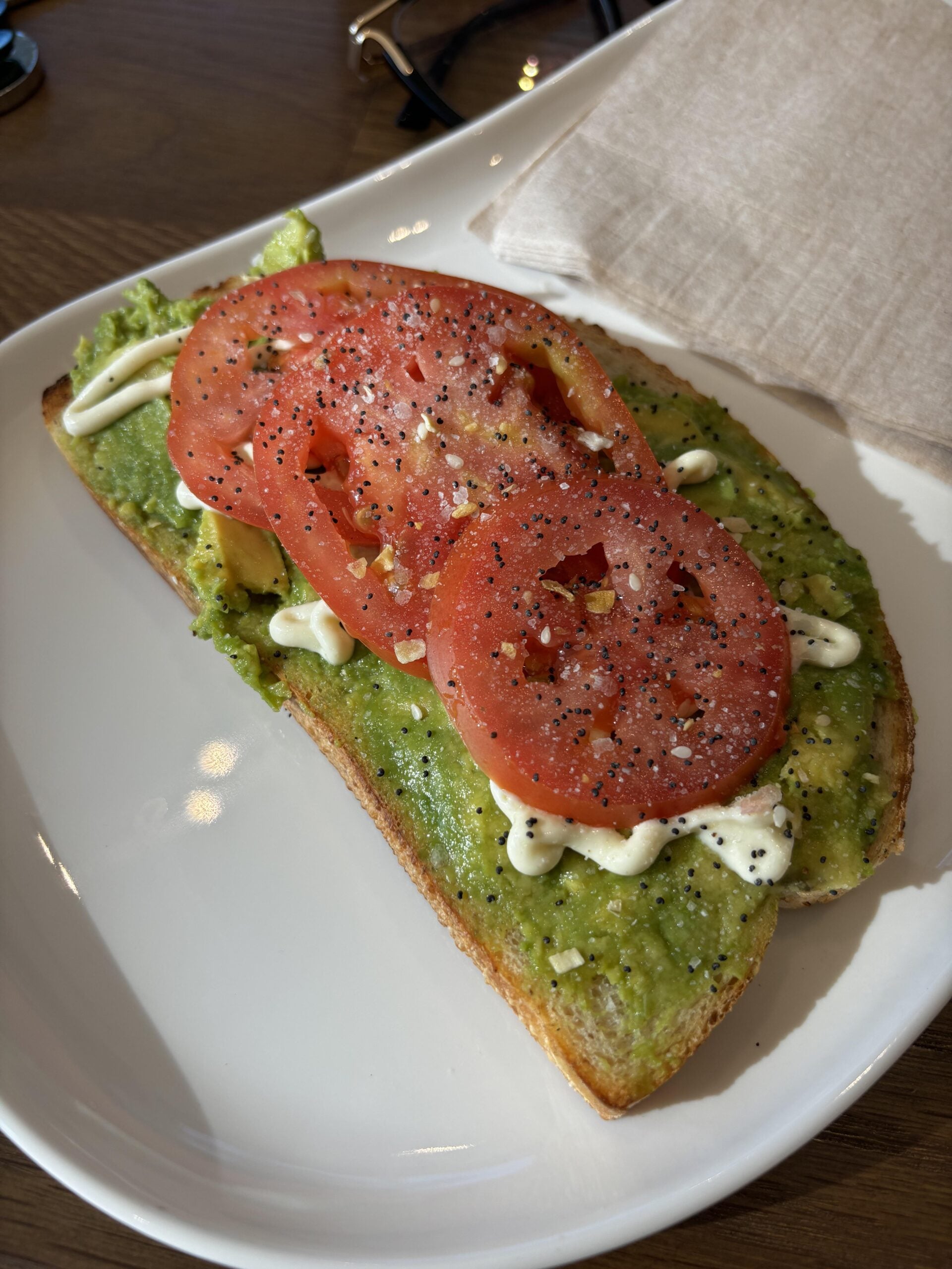 A slice of toast topped with mashed avocado, two slices of tomato, mayonnaise, and black pepper, served on a white plate with a beige napkin and dark glasses in the background.