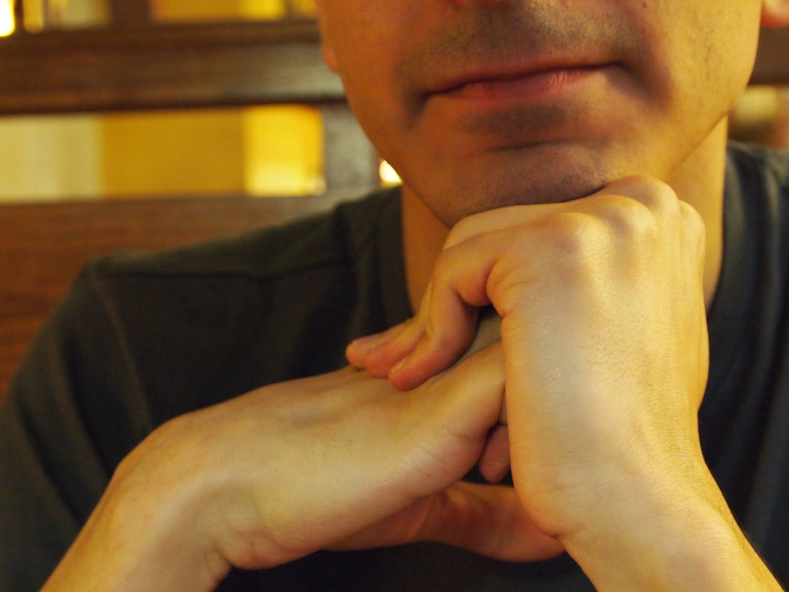 A person rests their chin on their clasped hands, sitting in front of a wooden background. Only the lower half of the face and hands are visible. The person wears a dark shirt.