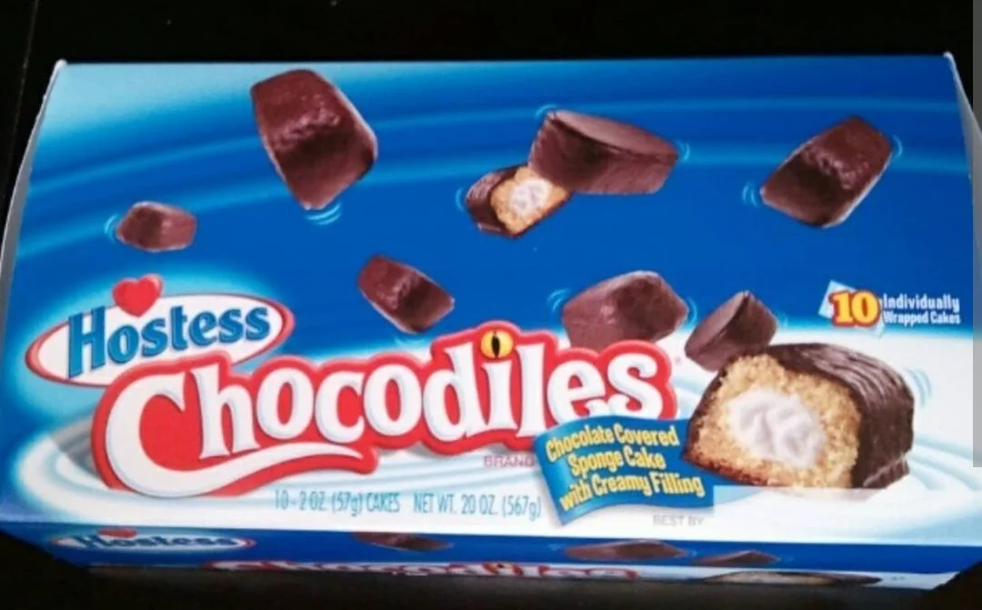 A blue box of Hostess Chocodiles, showing chocolate-covered sponge cakes with creamy filling. The front of the box displays cakes cut open to reveal the filling, and notes 10 individually wrapped cakes.