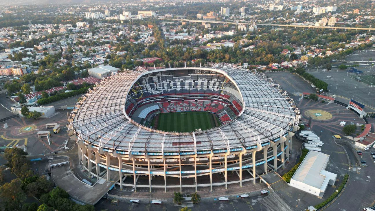 Aerial view of a large, circular soccer stadium with a green field and seating areas, surrounded by roads, buildings, and trees in an urban area.